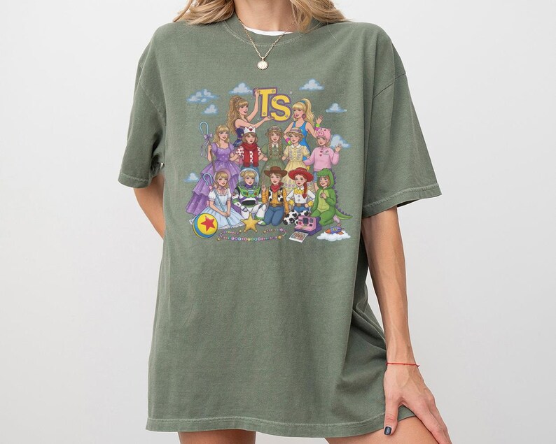 May include: An olive green short-sleeve t-shirt featuring a graphic print of various characters dressed in outfits inspired by Toy Story. The design includes the yellow text TS at the top, with characters arranged in a group against a blue sky background.