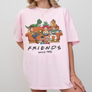 K&ouml;nnte beinhalten: Ein hellrosa T-Shirt mit einer Grafik verschiedener animierter Spielzeugfiguren, die auf einem orangefarbenen Sofa sitzen. Unter dem Sofa steht der Text FRIENDS SINCE 1995 in schwarzen Gro&szlig;buchstaben mit bunten Punkten zwischen den Buchstaben von FRIENDS.