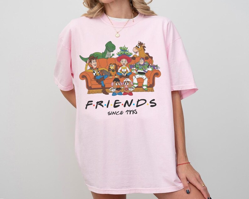 K&ouml;nnte beinhalten: Ein hellrosa T-Shirt mit einer Grafik verschiedener animierter Spielzeugfiguren, die auf einem orangefarbenen Sofa sitzen. Unter dem Sofa steht der Text FRIENDS SINCE 1995 in schwarzen Gro&szlig;buchstaben mit bunten Punkten zwischen den Buchstaben von FRIENDS.