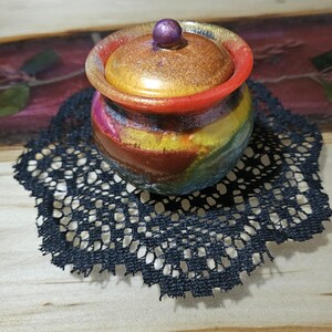 May include: A small, colorful ceramic jar with a lid. The jar has a rainbow-like pattern with shades of red, orange, yellow, green, blue, and purple. The lid is gold with a purple knob.