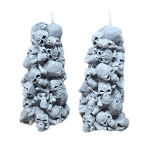 May include: Two gray skull-shaped candles. Each candle is a pillar of stacked skulls, with visible wicks at the top. The candles are set against a white background, highlighting their detailed, textured surfaces. A unique gothic home decor item.