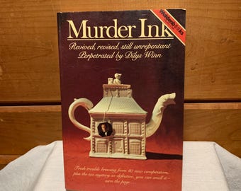 May include: A book cover with a red background and the title "Murder Ink" in white letters. The book is about a tea mystery and features a white teapot shaped like a house with a small figure of a person inside.