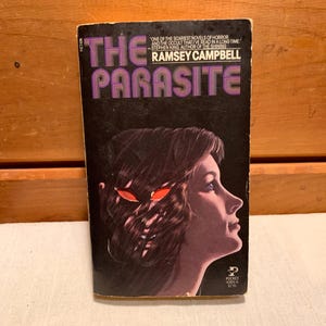 Op de afbeelding: Een paperback boekomslag met een zwarte achtergrond en een vrouwengezicht op de voorkant. De titel "The Parasite" staat in grote, vetgedrukte, paarse letters. De tekst "One of the scariest novels of horror and the occult that I've read in a long time" staat in witte letters. De tekst "Stephen King, author of The Shining" staat in witte letters. De tekst "Ramsey Campbell" staat in witte letters. De boekomslag heeft een Pocket Books logo in de rechterbenedenhoek.