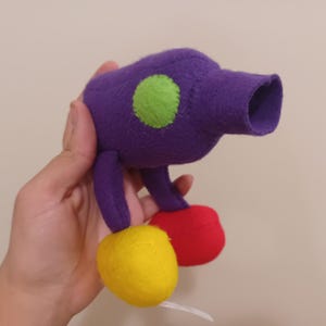 Könnte beinhalten: Lilafarbenes Filzspielzeug mit grünen und roten Filzdetails. Das Spielzeug hat einen langen, zylindrischen Körper mit abgerundetem Ende und zwei kurzen Beinen. Das Spielzeug wird in einer Hand gehalten.