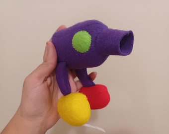 Può includere: Giocattolo di feltro viola con dettagli in feltro verde e rosso. Il giocattolo ha un corpo lungo e cilindrico con un'estremità arrotondata e due gambe corte. Il giocattolo è tenuto in una mano.