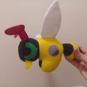 May include: A yellow and black felt bee plush toy with a red and green felt stinger. The bee has a white felt wing and a gray felt eye.