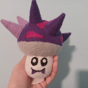May include: A handmade plush toy shaped like a mushroom with a cartoon face. The plush has a gray stem and a purple cap with pink accents and spikes. The face is white with black eyes and a smiling mouth.