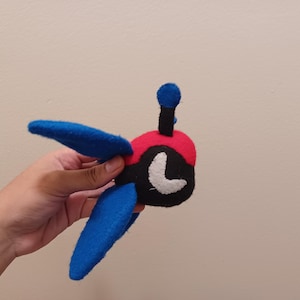 May include: A handmade plush toy featuring a red, black, and blue design. The toy has blue wings, a red head, and a black body with a white accent. It has a small antenna and is held in a hand.