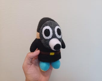 May include: A handmade plush toy resembling a cartoon character. It has a black body, a grey mask with large white eyes, and blue feet. A brown belt with a yellow buckle is also visible. The toy is held in a person's hand.
