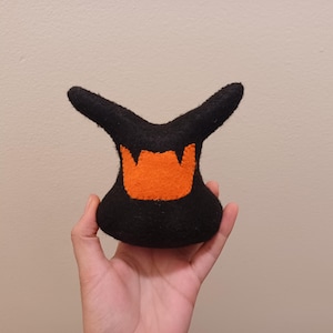 May include: A black felt plush toy with an orange flame-like design. The toy has two upward-pointing horns and a rounded base. The plush is held in a hand against a neutral background. The toy's design suggests a Halloween or fantasy theme.