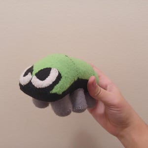 May include: A green and black plush toy with large eyes and four gray legs. The toy is shaped like a bug.