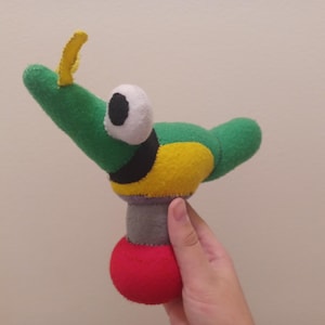 May include: A green, yellow, and black felt plush toy with a large white eye and a yellow antenna. The toy is shaped like a creature with a long neck and a round body. The toy is standing on a red base.