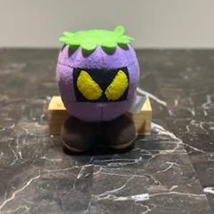 May include: Purple plush toy with a green leafy head, black eyes, and brown legs. The toy is sitting on a wooden block.