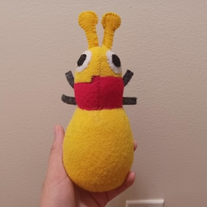 May include: A yellow felt plush toy with red and black accents. The toy has two antennas and large eyes.