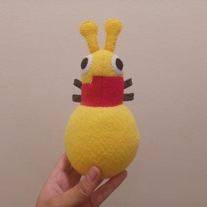 May include: A yellow felt plush toy shaped like a creature with two antennae, large white and black eyes, and a red patch on its chest. The toy features four small dark brown appendages on its sides and has a rounded, bulbous lower body section.
