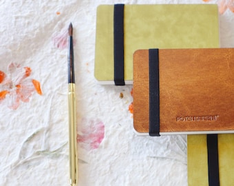 May include: Three blank sketchbooks with black elastic closures. The sketchbooks are stacked on top of each other. The top sketchbook is green, the middle sketchbook is brown, and the bottom sketchbook is yellow. The sketchbooks have the word "POTENTIALITY" printed on the front.