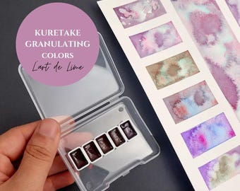 May include: A close-up of a KURETAKE GRANULATING COLORS L'art de Lime watercolor palette with six pans of dark brown paint. The palette is open and the pans are visible. The palette is sitting on a white surface.