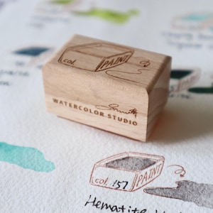 May include: A wooden stamp for watercolor paint, with the words "PAINT" and "Watercolor Studio" etched on it. The stamp is rectangular with a beveled edge. The background shows watercolor swatches and color names.