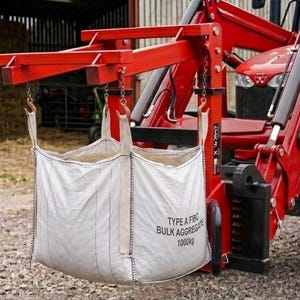 May include: A bright red tractor with a front-end loader attachment lifting a large white bulk bag. The bag is labeled TYPE A FIBC BULK AGGREGATE 1000kg and is suspended by four straps attached to metal hooks on the red lifting frame of the tractor.