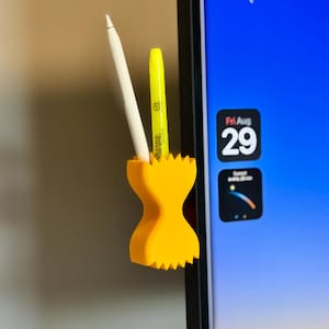 Könnte beinhalten: Ein leuchtend gelber Stifthalter in Sanduhrform, an einem Computermonitor befestigt. Er enthält einen weißen Stift und einen gelben Sharpie-Marker. Das Design hat einen gezackten Rand.