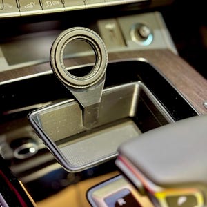May include: Close-up of a black gear shift knob with a silver ring around the top. The knob is in a car's center console with a black and silver gear selector.
