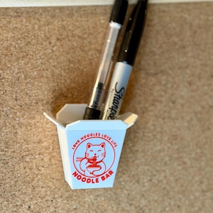 Könnte beinhalten: Ein kleiner weißer Imbissbehälter mit rotem Text und einer Katzenzeichnung, der zwei Stifte enthält. Der Behälter trägt die Aufschrift "Love Noodles Love Life" und "Noodle Bar". Ein Stift ist ein schwarzer Sharpie, der andere ist ein durchsichtiger Stift mit schwarzer Tinte.