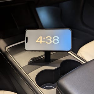 May include: A smartphone displaying the time 4:38 is mounted on a black stand inside a car. The phone is centered on a console with cup holders. The interior features black and cream-colored leather.