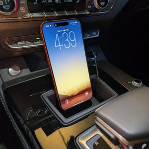 May include: A smartphone with an orange frame and a digital display showing the time 4:39, date Sun 6, and temperature 69 degrees, positioned upright within a black rectangular car console storage tray located near the gear shift lever.