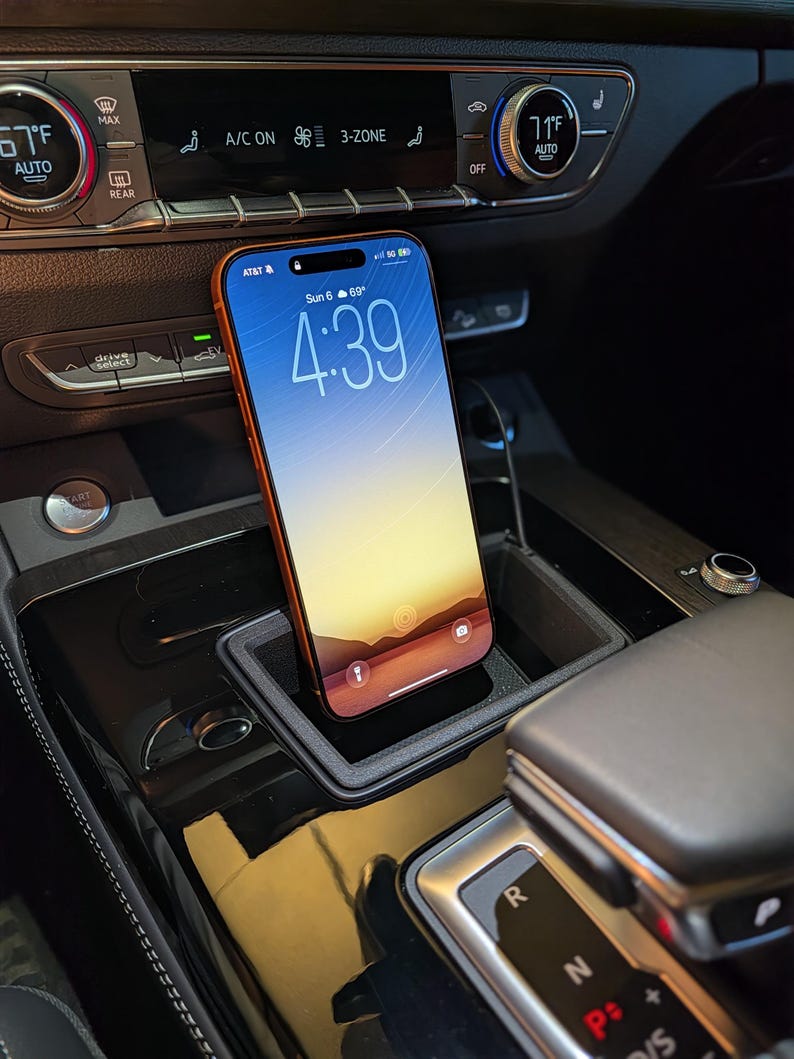 May include: A smartphone with an orange frame and a digital display showing the time 4:39, date Sun 6, and temperature 69 degrees, positioned upright within a black rectangular car console storage tray located near the gear shift lever.