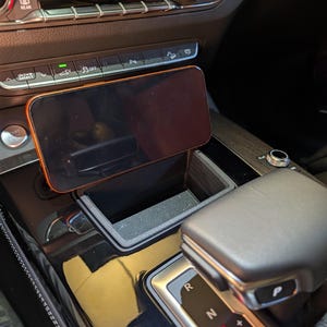 May include: A black smartphone mounted in a custom-fit plastic holder positioned within the centre console storage compartment of a vehicle, located directly in front of the climate control panel and gear shift selector.