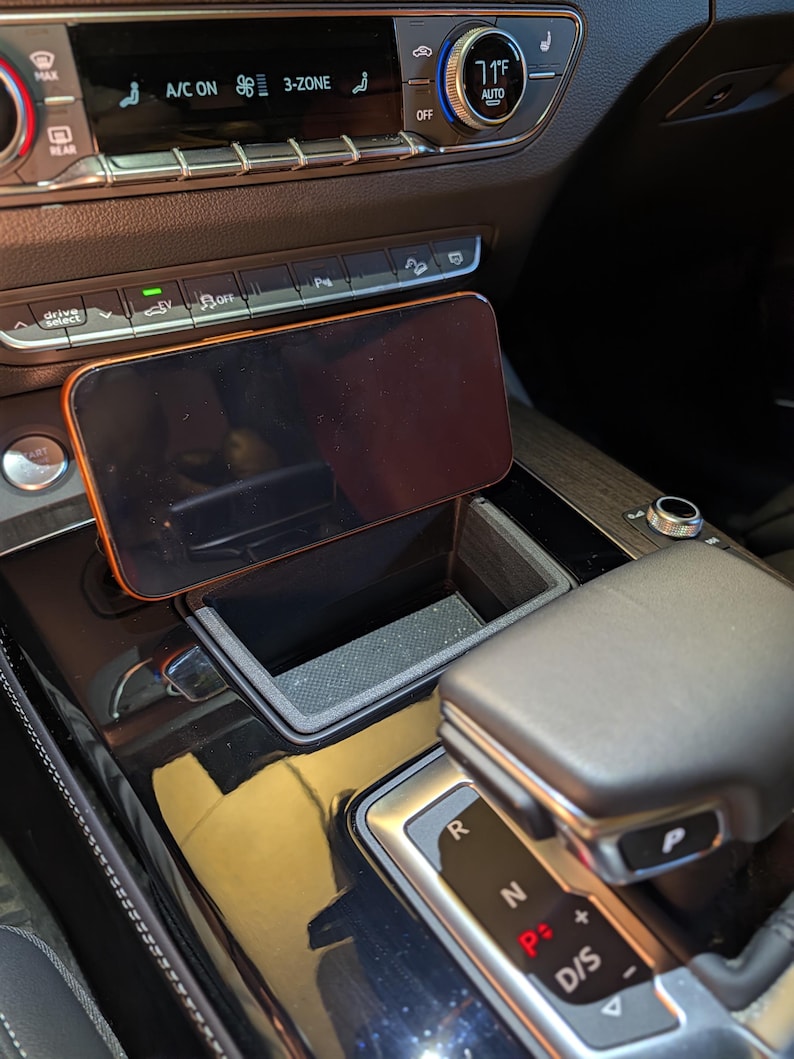 May include: A black smartphone mounted in a custom-fit plastic holder positioned within the centre console storage compartment of a vehicle, located directly in front of the climate control panel and gear shift selector.