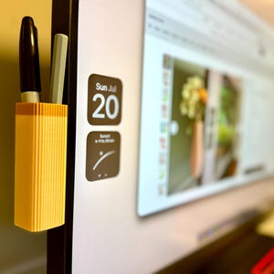 May include: A yellow and orange pen holder attached to a computer monitor, holding a black marker and a gray pen. The monitor displays a webpage with a blurred image. The date "Sun Jul 20" is visible on the screen.