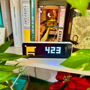 Pode incluir: Um relógio digital branco com um ícone de carrinho de compras amarelo e o número 423 exibido em um fundo preto. O relógio está sentado em uma prateleira com livros e uma casa de madeira em miniatura atrás dele.