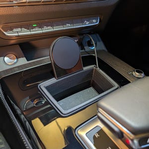 May include: A black, 3D-printed rectangular storage tray designed for a vehicle centre console, featuring an integrated circular magnetic mount for a mobile device. The tray has a textured finish and fits into the existing console space.