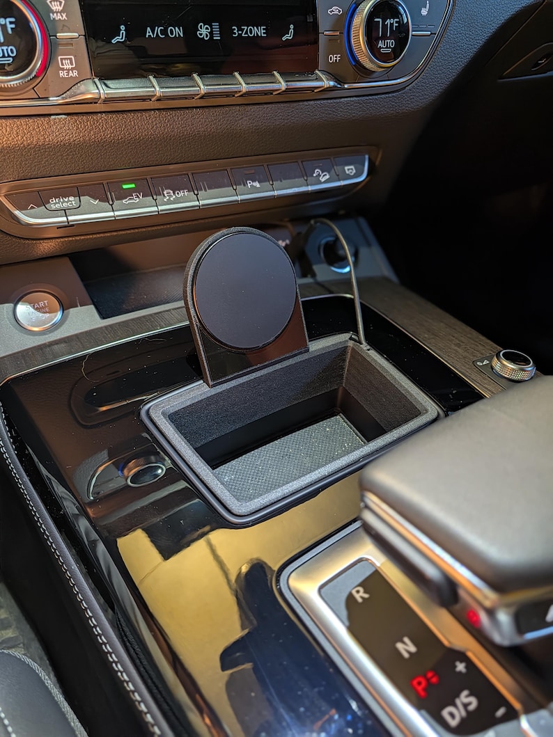 May include: A black, 3D-printed rectangular storage tray designed for a vehicle centre console, featuring an integrated circular magnetic mount for a mobile device. The tray has a textured finish and fits into the existing console space.