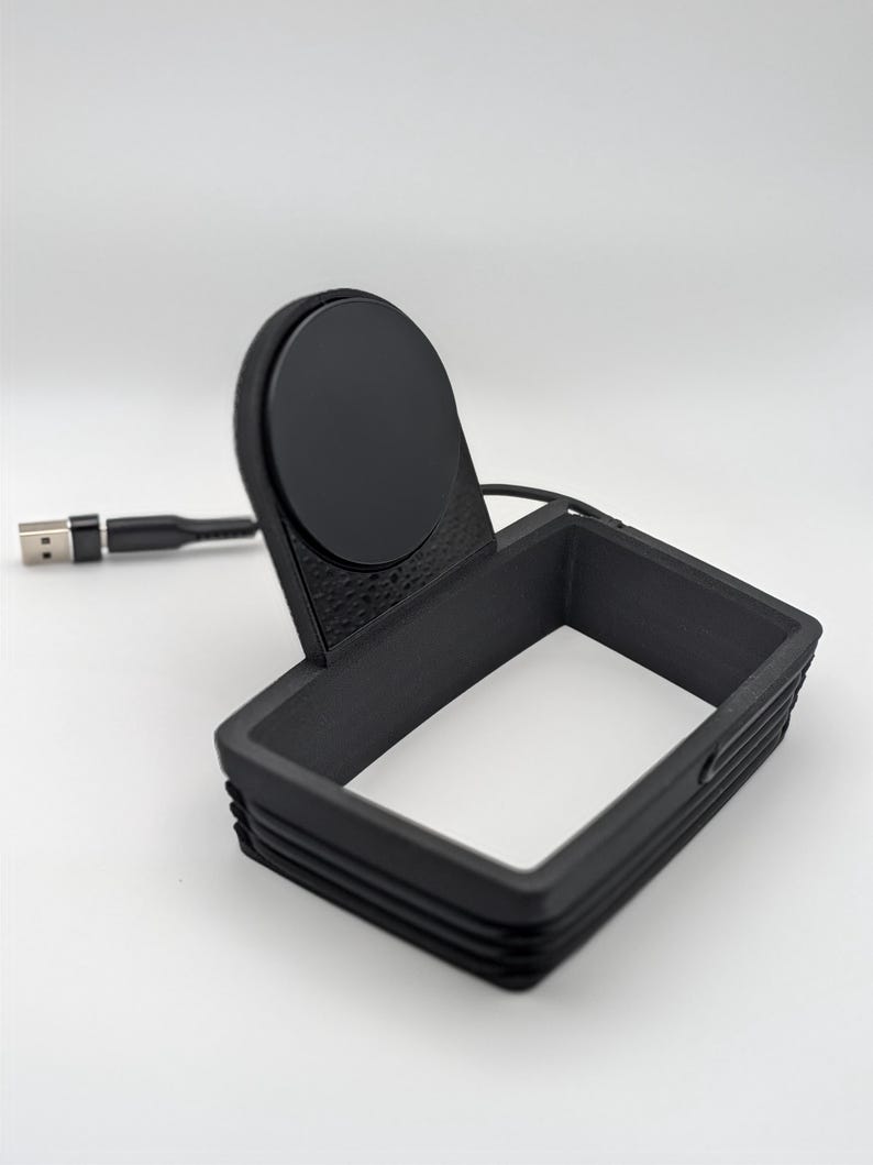 May include: A black matte charging stand featuring a rectangular frame base and a circular magnetic charging disc attached to an upright support. A black USB cable extends from the back of the unit, ending in a standard USB-A connector plug.