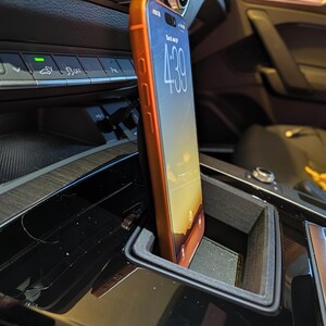 May include: A smartphone with an orange case is positioned vertically in a black plastic holder mounted within the centre console of a vehicle. The phone screen displays the time 4:39 and a sunset wallpaper, with a charging cable connected at the base.