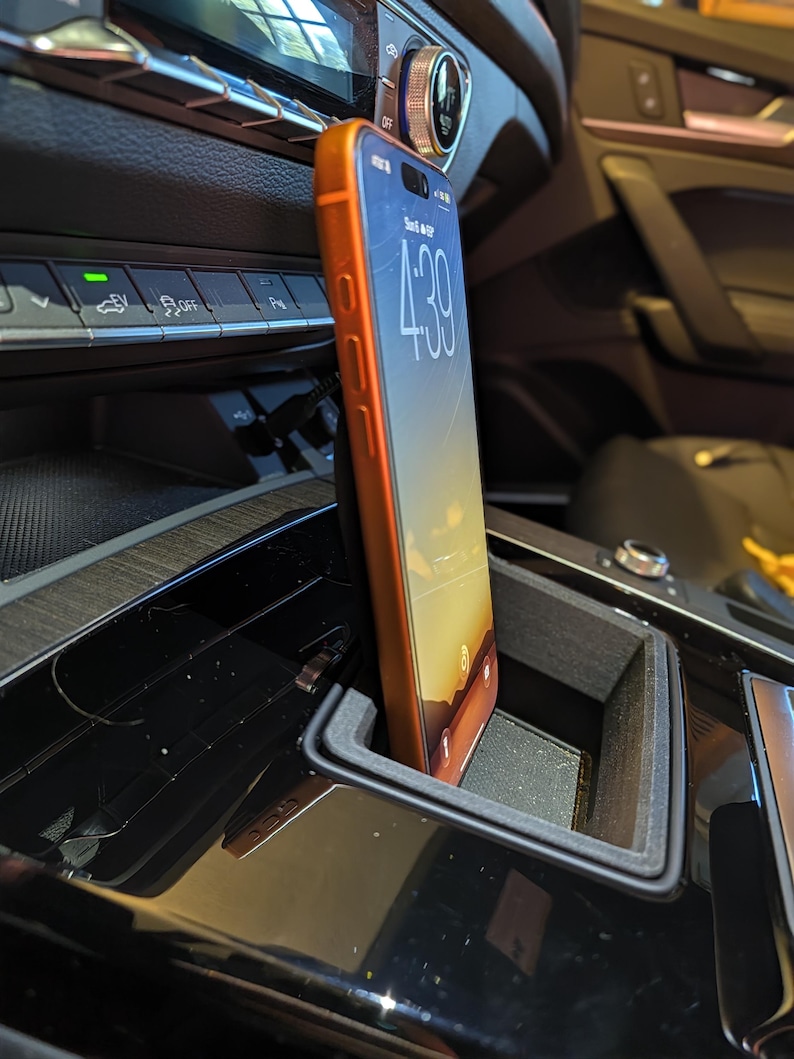 May include: A smartphone with an orange case is positioned vertically in a black plastic holder mounted within the centre console of a vehicle. The phone screen displays the time 4:39 and a sunset wallpaper, with a charging cable connected at the base.