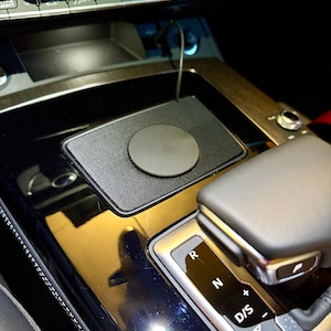 以下が含まれることがあります： 黒色のワイヤレス電話充電器。円形の充電パッドが車のコンソールに設置されています。充電器は長方形で、光沢のある黒い表面にあります。 ギアレバーが見えます。