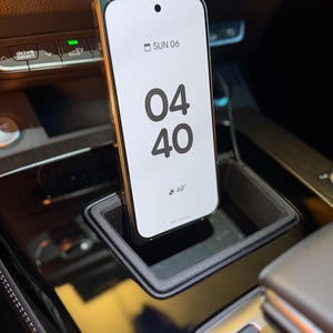 May include: A smartphone with a white screen displaying the time 04:40 and the date SUN 06, positioned vertically within a black rectangular car console storage slot. The phone screen also shows a weather icon with 68 degrees and 20% charging.