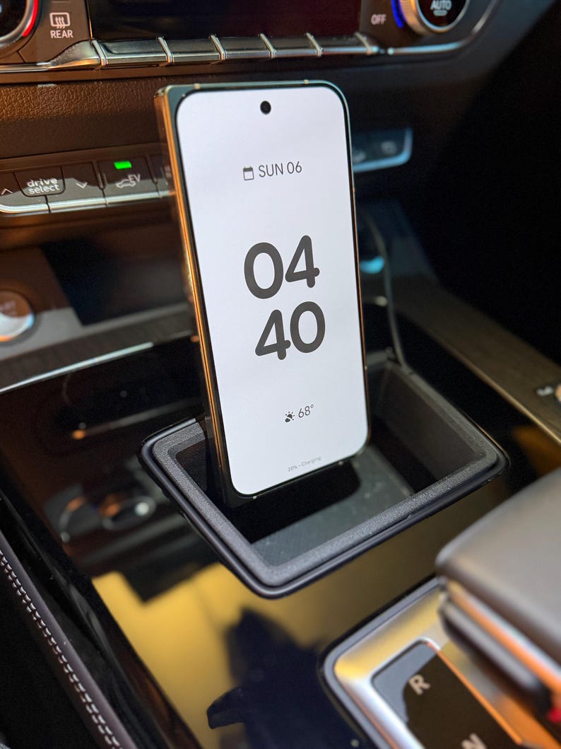May include: A smartphone with a white screen displaying the time 04:40 and the date SUN 06, positioned vertically within a black rectangular car console storage slot. The phone screen also shows a weather icon with 68 degrees and 20% charging.