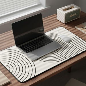 May include: A laptop, mouse, and speaker rest on a wooden desk. A large, rectangular mousepad with a black and white geometric design is beneath the laptop and mouse. A notebook and pen are also on the desk.