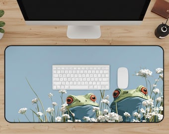 May include: A rectangular desk mat featuring a light blue background with a whimsical illustration of two green frogs with red eyes amidst white flowers. A white keyboard and mouse are on the mat. The mat is surrounded by a black border.