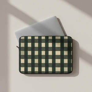 Puede incluir: Una funda para portátil con un diseño de cuadros verdes y crema. La funda está abierta, revelando un portátil plateado. La funda tiene una cremallera negra y un borde negro. El patrón de cuadros es clásico.