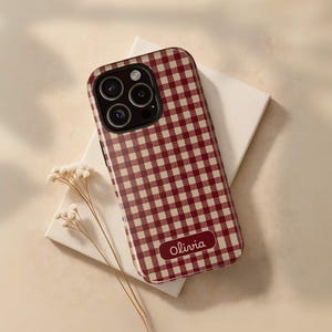 Puede incluir: Funda de teléfono con estampado de cuadros vichy burdeos y crema con una cámara de tres lentes. La funda tiene el nombre "Olivia" impreso. La funda del teléfono está sobre una superficie rectangular blanca con flores secas.