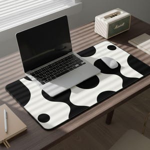 May include: A black and white patterned desk mat with a laptop, mouse, and speaker on a wooden desk. The mat features a repeating pattern of black circles and curved shapes. A notebook and pen are also on the desk.