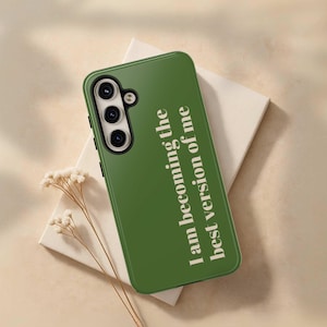 May include: Green phone case with the text "I am becoming the best version of me" in white. The phone case has a camera module with three lenses. The case is resting on a white surface with a beige background.