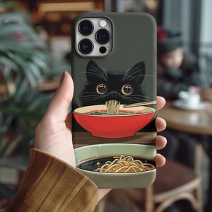 Puede incluir: Un gato negro con ojos verdes está comiendo fideos de un tazón rojo. El gato está en una funda de teléfono con fondo negro y una lente de cámara. La funda del teléfono también tiene un tazón de fideos.