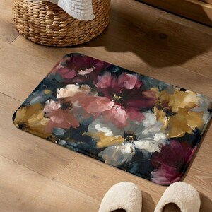 May include: A rectangular bath mat with a floral design in shades of pink, white, yellow, and burgundy. The flowers are painted on a dark background. The mat is on a wooden floor, near a wicker basket and a pair of white slippers.