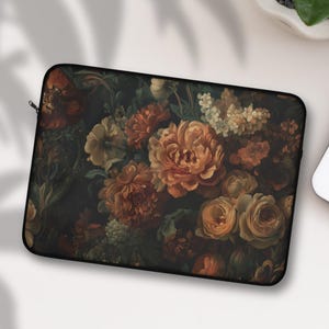 Op de afbeelding: Een zwarte laptoptas met een vintage bloemenpatroon. Het ontwerp toont een donkere achtergrond met bloemen in oranje, gele en crèmetinten. De hoes heeft een ritssluiting en is ontworpen om een laptop te beschermen.